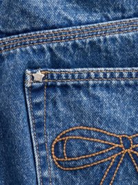 Blå denimstoff med et stort, brun sydd design som ligner en sløyfe. Inkluderer et metallmerke med stjerneform og synlig søm.