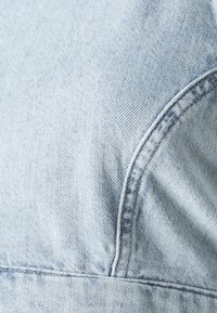 Tessuto in denim blu chiaro con una texture liscia, cuciture visibili e un motivo leggermente sbiadito vicino ai bordi curvi.