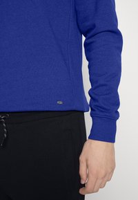 Maglietta a maniche lunghe blu royal realizzata in tessuto morbido, con scollatura e polsini a coste, e un piccolo logo metallico in fondo.