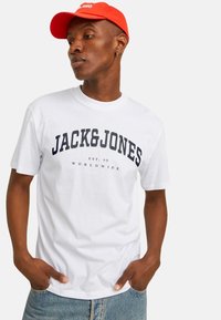 Hvid bomulds-t-shirt med marineblå "JACK&JONES" tekst, der har en klassisk rund hals. Sammen med lyseblå denimjeans og en rød cap.
