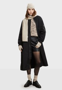 Lang sort yderjakke over en lys sweater, sort lædernederdel og sorte strømpebukser. Tilbehør inkluderer en beige hue og tørklæde.