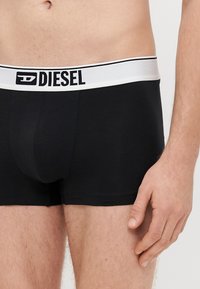 Melni kokvilnas bokseri ar baltu elastīgu jostu, uz kuras ir melns uzraksts "DIESEL". Gluda tekstūra, pieguļošs dizains, vidēja garuma līdz augšstilbam.