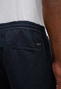 Pantaloni sportivi blu scuro con elastico in vita, tasca laterale e un'etichetta cucita sulla tasca posteriore. Il tessuto appare liscio con una texture minima.