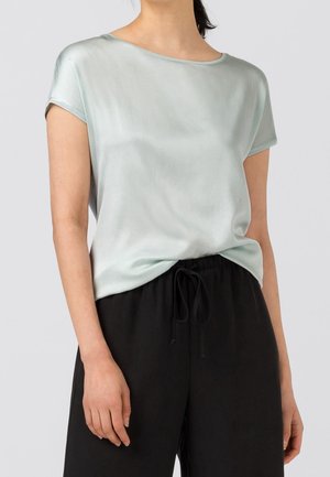 Mujer que lleva una blusa de satén azul claro de manga corta metida dentro de unos pantalones negros con cordón, sobre un fondo liso.