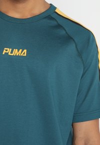 PUMA turkos kortärmad t-shirt med rund hals; har gula och svarta ränder längs axlarna och en logotyp på bröstet.
