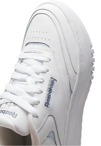 Sneaker en cuir blanc avec une surface texturée, languette rembourrée et lacets blancs. Présente un accent bleu et un logo Reebok sur le côté.