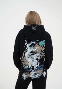 Completo felpa con cappuccio e pantaloni tuta neri che presenta una grande grafica di drago con accenti floreali, dettagli ricamati sul cappuccio e una texture morbida.