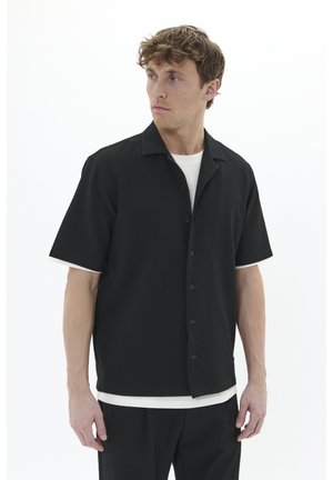 Kortærmet sort skjorte med et struktureret mønster, knaplukning foran og åben krave, båret over en hvid t-shirt med rund hals.