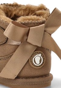 Zapato para bebé texturizado marrón con forro suave de piel, lazo de satén y emblema circular con una huella en el lateral.