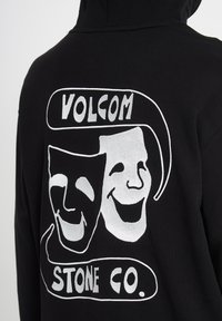 Sweat à capuche noir avec des masques théâtraux blancs de comédie et de tragédie et le texte "Volcom Stone Co." au dos.