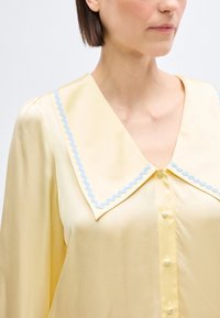 Γυναίκα που φοράει ανοιχτό κίτρινο μπλουζάκι με μεγάλο μυτερό γιακά διακοσμημένο με γαλάζια σκοινί και κουμπιά καλυμμένα με ύφασμα στη μπροστινή πλευρά.