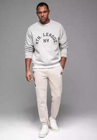 Šedá mikina s černým potiskem "ATH. LEAGUE NY", spárovaná s světle béžovými tepláky s bočními kapsami a černými akcenty, nošená s bílými teniskami.