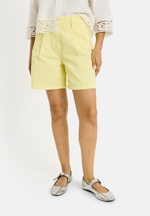 Personne portant un short jaune pâle au genou, un haut blanc avec des manches en dentelle et des chaussures plates Mary Jane argentées, debout sur un fond blanc.