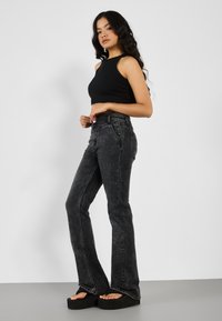 Jeans acampanados negros con un lavado desgastado, cintura alta, bolsillos frontales y sandalias de plataforma negras. La modelo lleva una blusa sin mangas negra ajustada.