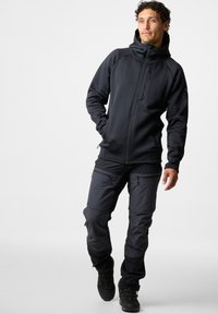 Felpa nera con zip e pantaloni scuri con molte tasche, realizzati in un materiale morbido ed elastico. Presenta un cappuccio e minimi dettagli in metallo.