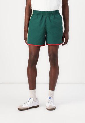 Persona vistiendo pantalones cortos deportivos verde oscuro con ribete rojo, calcetines blancos y zapatillas Adidas blancas, de pie contra un fondo blanco.