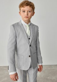 Completo grigio con tessuto strutturato, due bottoni e tasche. Abbinato a una camicia bianca e una cravatta verde chiaro a fantasia. Vestibilità sartoriale.