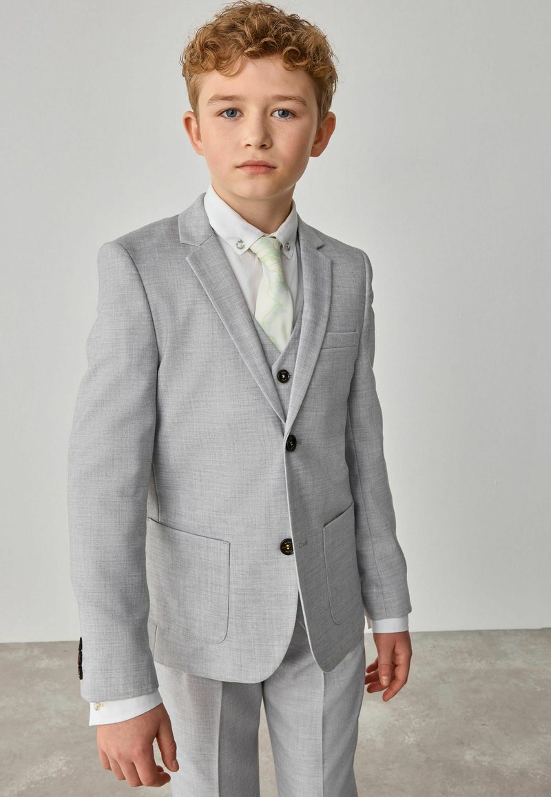 Completo grigio con tessuto strutturato, due bottoni e tasche. Abbinato a una camicia bianca e una cravatta verde chiaro a fantasia. Vestibilità sartoriale.