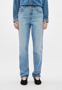 Svetlo modri kavbojki z ravnim krojem, ki imajo sprednje žepe in rahlo zbledel videz. Obuti z črnimi natikači.