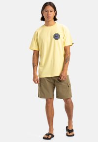 Camiseta de algodón amarilla con un gráfico circular en el pecho, combinada con pantalones cortos de carga en color caqui. Chanclas negras completan el atuendo casual.
