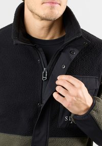 Giacca in fleece nera e verde oliva con chiusura a zip, bottoni a pressione, tessuto testurizzato e tasca sul petto con dettaglio del logo.