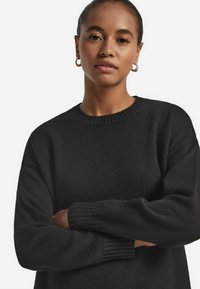 Schwarzer Strickpullover mit rundem Halsausschnitt, gerippten Bündchen und lockerer Passform. Hergestellt aus weichem Material mit einer strukturierten Oberfläche.