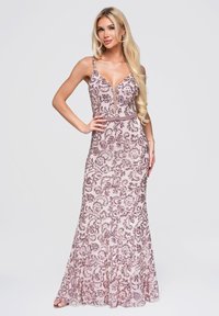 Femme aux longs cheveux blonds portant une robe rose sans manches longue jusqu'au sol avec des motifs floraux foncés, debout avec une main sur la taille.