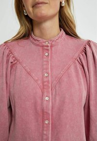 Rosa kordflöjls skjorta med hög krage, knappstängning framtill och plisserade puffärmar. Har snedställd ok och kontrasterande sömmar.