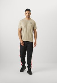 Beige short-sleeve shirt with a gray logo, paired with black pants featuring red and white side stripes and black sneakers.