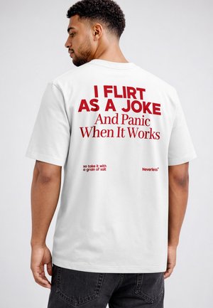 Mann trägt ein weißes T-Shirt mit rotem Text auf dem Rücken „Ich flirte aus Spaß und gerate in Panik, wenn es funktioniert“ und steht vor einem einfarbigen Hintergrund.