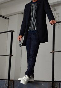 Navy wool coat over a patterned knit sweater, paired with navy checkered pants and white sneakers. Person holds glasses in one hand.