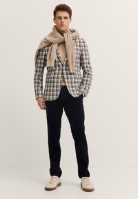 Herren kariertes Blazer in Beige und Blau, ergänzt durch einen beigen Pullover und marineblaue Hosen. Die Schuhe sind cremefarbene Sneakers mit einer beigen Sohle.