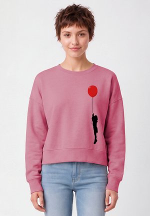 Femme portant un sweat-shirt rose avec une silhouette noire d'une fille tenant un ballon rouge imprimé sur le devant, associé à un jean bleu clair.