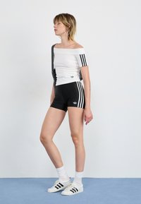 Balts off-the-shoulder tops ar melnām trīslinijām, apvienots ar melniem šortiem, kam ir līdzīgas strīpas, un baltām kedām ar melnām strīpām.