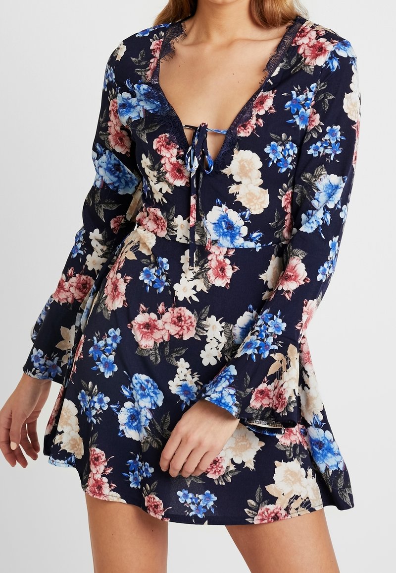 Kukallinen mekko, jossa on tummansininen tausta ja vaaleanpunaisia, sinisiä ja valkoisia kukkia. Pitkät hihat, V-pääntie pitsireunalla, ja nauhakiinnitys edessä.