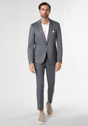 Man loopt vooruit, draagt een grijs pak met een wit overhemd, beige sneakers en een neongroene pochet op een witte achtergrond.