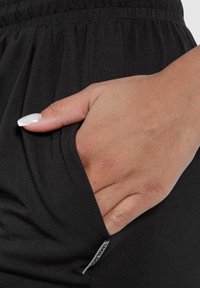Zwarte stoffen broek met een elastische tailleband en zijzak. Close-up van een hand die in de zak rust, waarbij de soepele textuur en afwerking zichtbaar zijn.