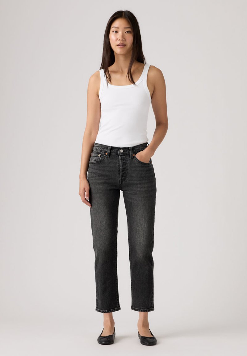 Black denim jeans with a straight cut, high waist, and four pockets. Worn with a fitted white tank top and flat black shoes, against a neutral background.