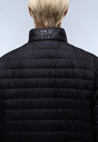 Personne aux cheveux blonds courts portant une veste matelassée noire avec un col haut et un logo partiellement visible.