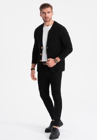 Černý pletený kardigan s dřevěnými knoflíky, kombinovaný s bílým tričkem, černými kalhotami a černými šněrovacími botami. Jednoduchý, minimalistický design.