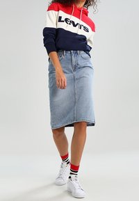 Sweat à capuche Levi's rouge, blanc et marine avec logo; associé à une jupe en denim bleu clair; baskets blanches et chaussettes rayées.