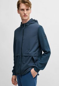 Marineblau gehäuften Jacke, die glattes Gewebe mit strukturierten Strickärmeln kombiniert. Sie verfügt über einen Frontreißverschluss, zwei Seitentaschen und ein Logo-Patch am Ärmel.