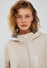 Light beige leather jacket with a hood, silver zipper, and contrasting black detailing at the collar. Smooth texture and minimalist design.