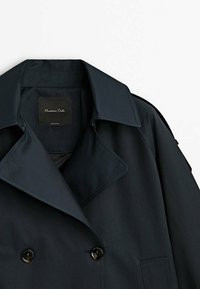 Granatowy podwójnie zapinany trencz z dużymi klapami, czarnymi guzikami i gładkim, teksturowanym materiałem. Metka widoczna wewnątrz kołnierza.