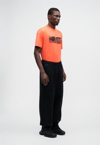 Tricou grafic portocaliu cu un design imprimat pe față, asociat cu pantaloni de trening negri largi și adidași negri.