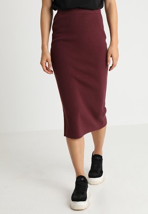Femme portant une jupe midi en maille bordeaux taille haute, des baskets noires et un top noir, marchant sur un fond blanc uni.