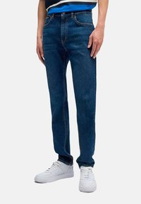 Jeans de mezclilla azul oscuro con un diseño de pierna recta, que cuentan con bolsillos estándar y detalles de costura, combinados con zapatillas blancas.