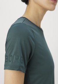 Tmavě zelené sportovní tričko vyrobené ze měkké, pružné tkaniny. Má kulatý výstřih a na levém rukávu je vytištěné logo "BORG".