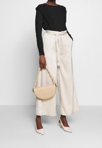 Sac à main beige texturé à forme courbée et poignée tressée, associé à un haut noir et un pantalon large crème.