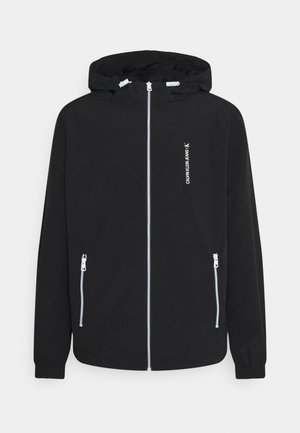 Czarna kurtka z kapturem, z białym zamkiem błyskawicznym z przodu, kieszeniami zapinanymi na zamek po bokach, elastycznymi mankietami oraz pionowym logo "Calvin Klein Jeans" na piersi.